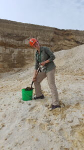 Person wearing hard hat standing in sandy area digging with a shovel. 